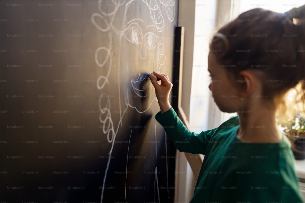 A little girl drawing with chalks on blackboard wall indoors in playroom.