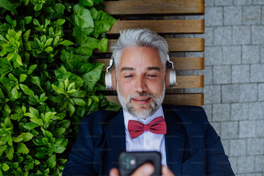 Um empresário maduro desfrutando de música em fones de ouvido relaxando no banco no parque da cidade durante o intervalo no trabalho.