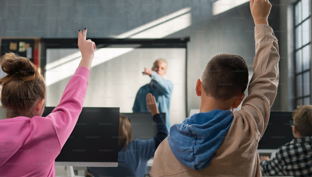 Una vista trasera de los niños de la escuela levantando las manos durante las pruebas de computadora en la escuela.