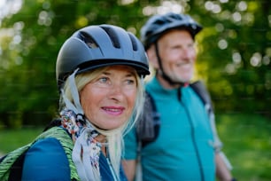 An active senior couple riding bicycles at summer park, woman with bicycle helmet, healthy lifestyle concept.