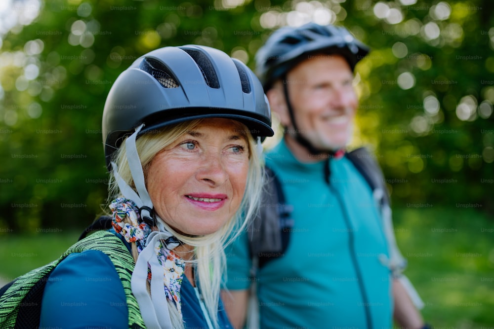An active senior couple riding bicycles at summer park, woman with bicycle helmet, healthy lifestyle concept.