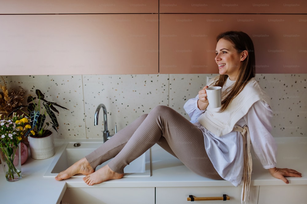 Una joven alegre sosteniendo una taza de café mientras está sentada en el mostrador de la cocina