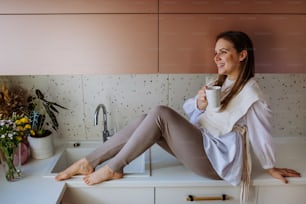 A cheerful young woman holding coffee cup while sitting on the kitchen counter