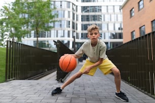 Cheerful caucasian boy driblling with basketball ball in public city park, looking at camera.