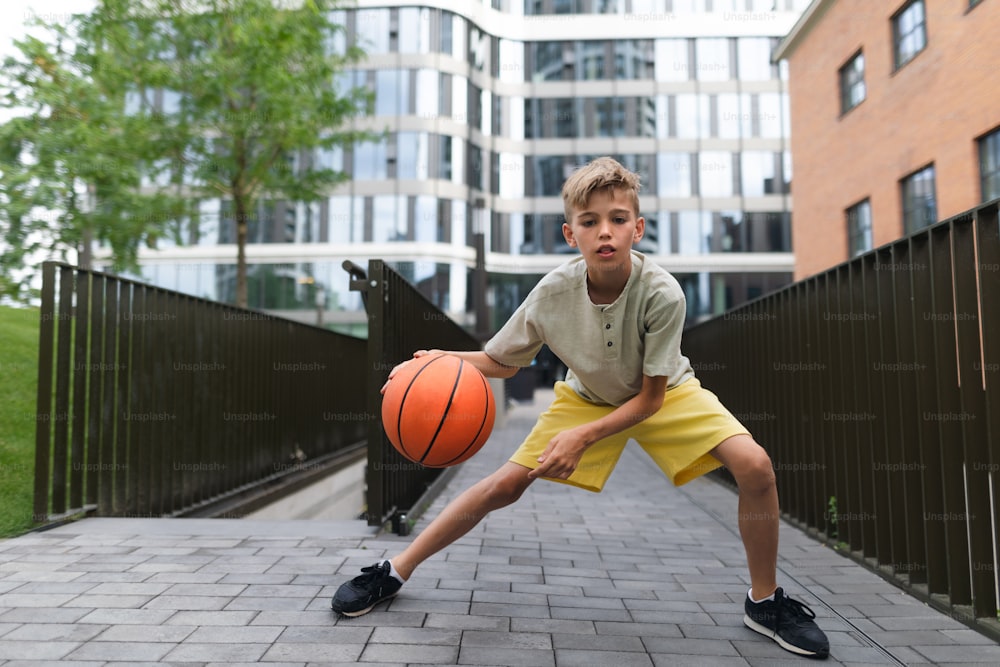 Alegre niño caucásico driblando con pelota de baloncesto en el parque público de la ciudad, mirando a la cámara.