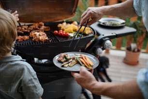 An unrecognizable father with little son grilling ribs and vegetable on grill during family summer garden party, close-up