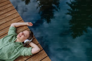 A rlaxed woman wearing headphones listening to music lying on a pier by natureal lake in summer
