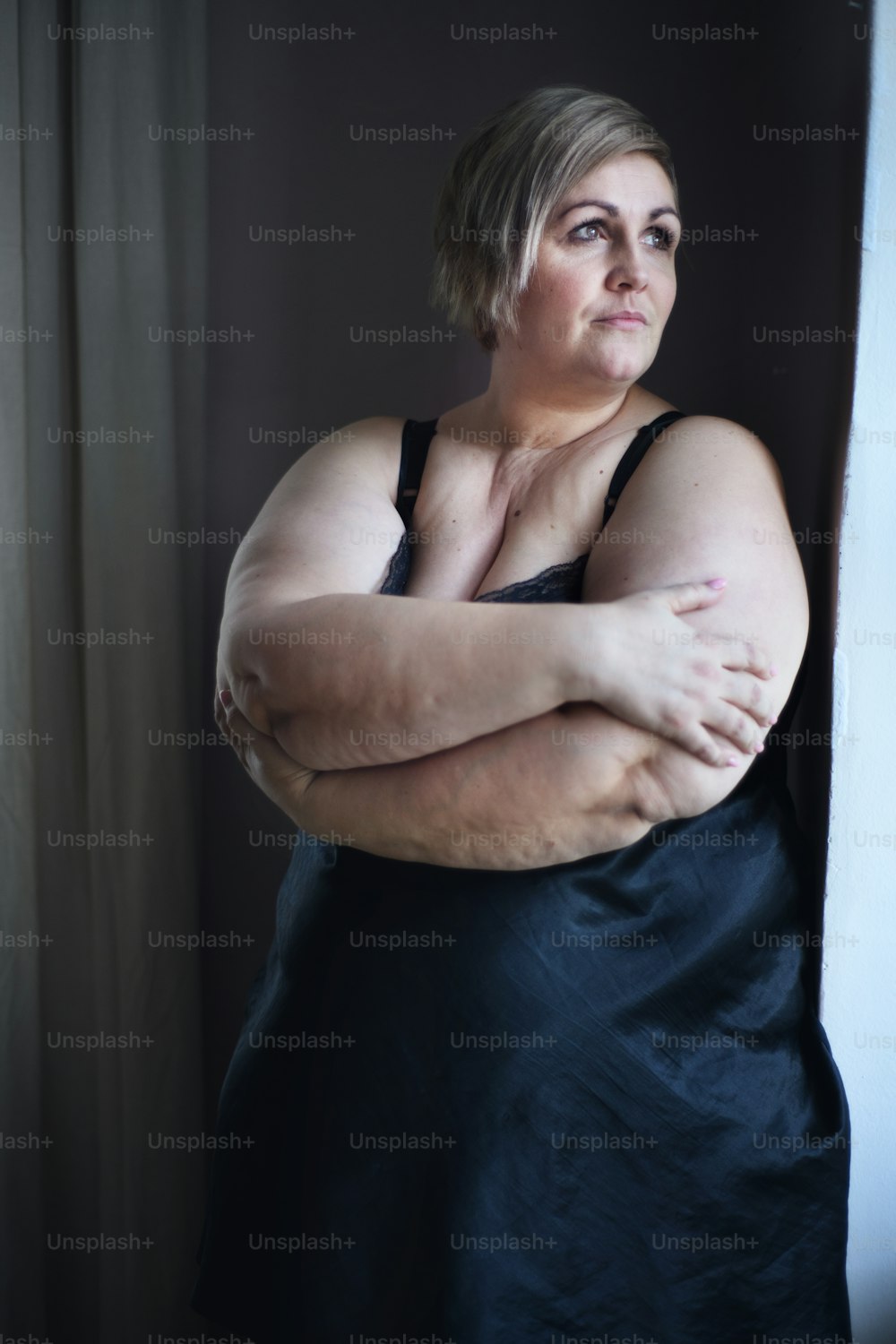 Una mujer gorda, solitaria y deprimida de pie mirando a través de la ventana de su casa.