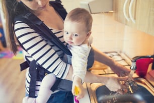 Young mother washing up dishes with her little daughter that she has in a baby carrier