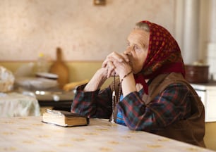 Mulher muito velha usando lenço na cabeça está rezando em sua cozinha estilo país