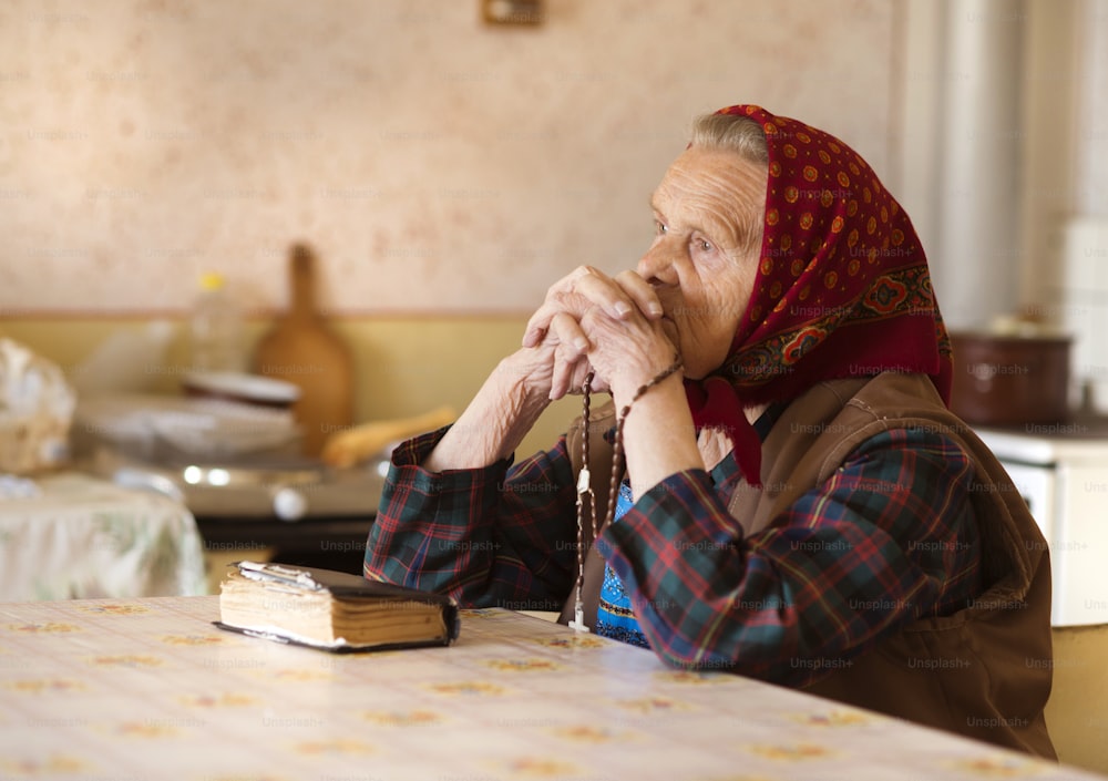 Una mujer muy anciana con un pañuelo en la cabeza está rezando en su cocina de estilo rústico