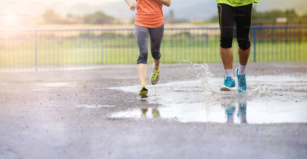 Giovane coppia che corre sul campo sportivo asfaltato in tempo piovoso. Dettagli di gambe e scarpe sportive che schizzano nelle pozzanghere.