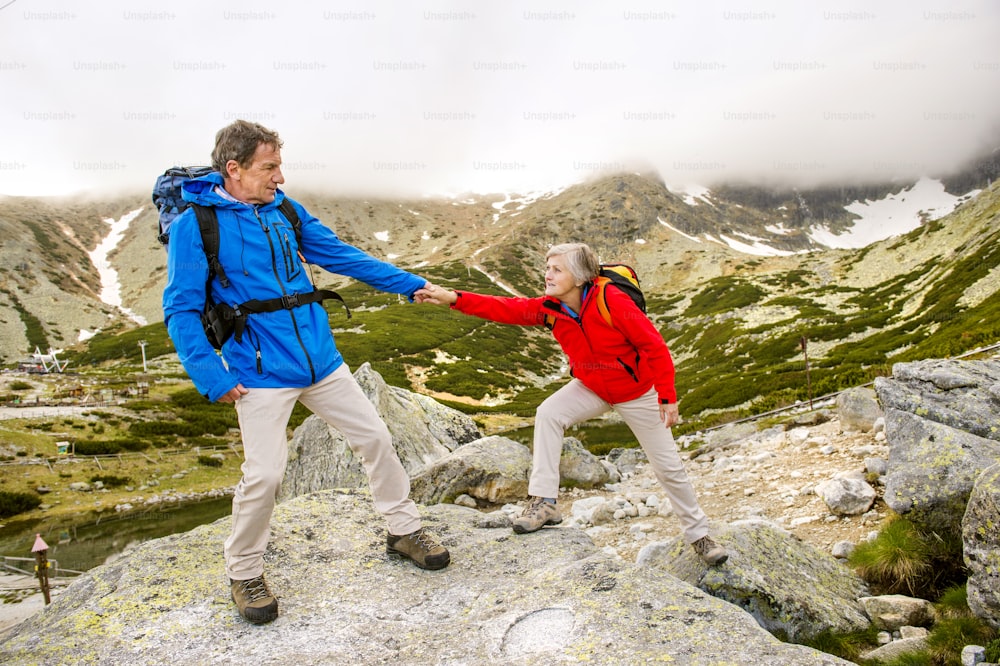 Casal de turistas sênior caminhando, o homem está ajudando a mulher a chegar à rocha