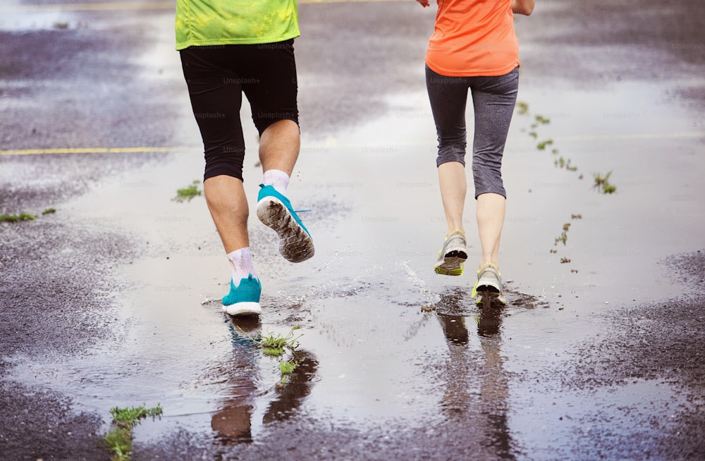 Jovem casal correndo em campo de esportes de asfalto em tempo chuvoso. Detalhes de pernas e sapatos esportivos espirrando em poças.