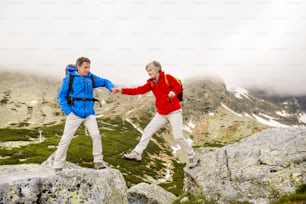Senior tourist couple hiking, man is helping woman to get to the rock