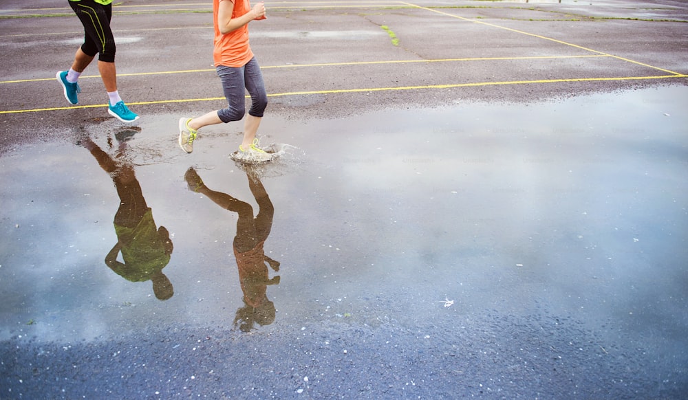 Jeune couple courant sur un terrain de sport asphalté par temps de pluie. Détails des jambes et des chaussures de sport éclaboussant dans les flaques d’eau.