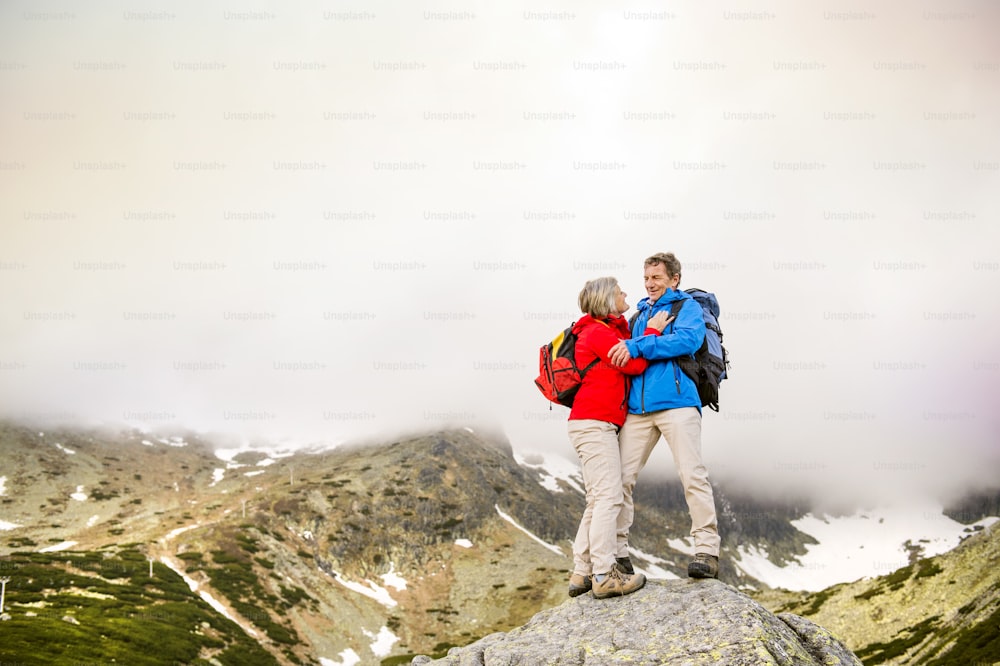 Senior tourist couple hiking, man is hugging womanon the rock