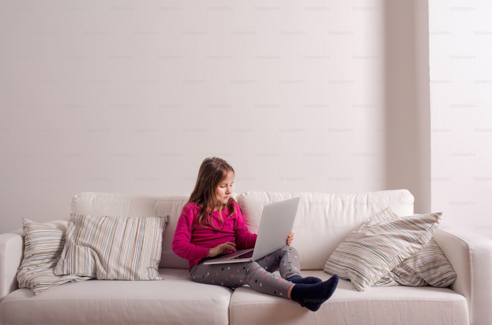 Little girl sitting on sofa with a laptop computer at home. Happy child playing indoors using PC, copy space