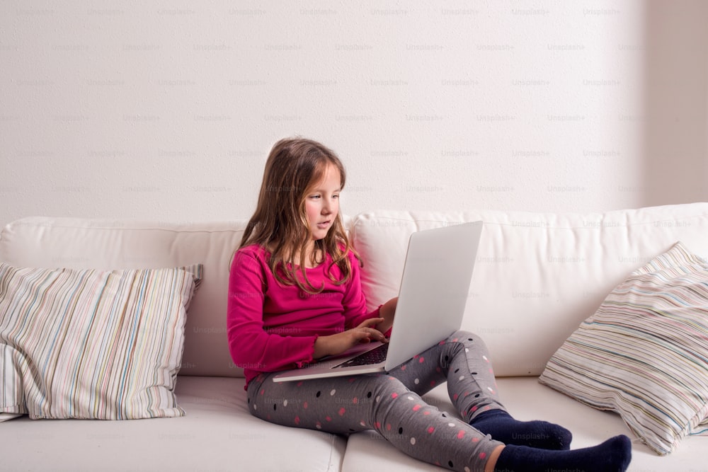 Little girl sitting on sofa with a laptop computer at home. Happy child playing indoors using PC.