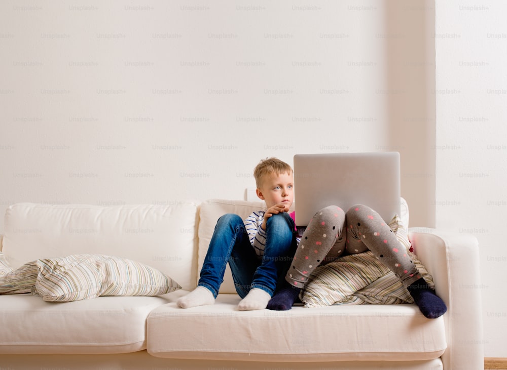 Petite fille et garçon assis sur un canapé avec un ordinateur portable à la maison. Des enfants heureux jouant à l’intérieur à l’aide d’un PC.