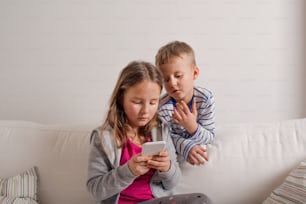 Little girl and boy sitting on sofa with a smart phone at home. Happy children playing indoors, copy space