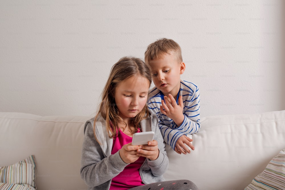 Little girl and boy sitting on sofa with a smart phone at home. Happy children playing indoors, copy space