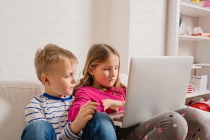 Petite fille et garçon assis sur un canapé avec un ordinateur portable à la maison. Des enfants heureux jouant à l’intérieur à l’aide d’un PC.