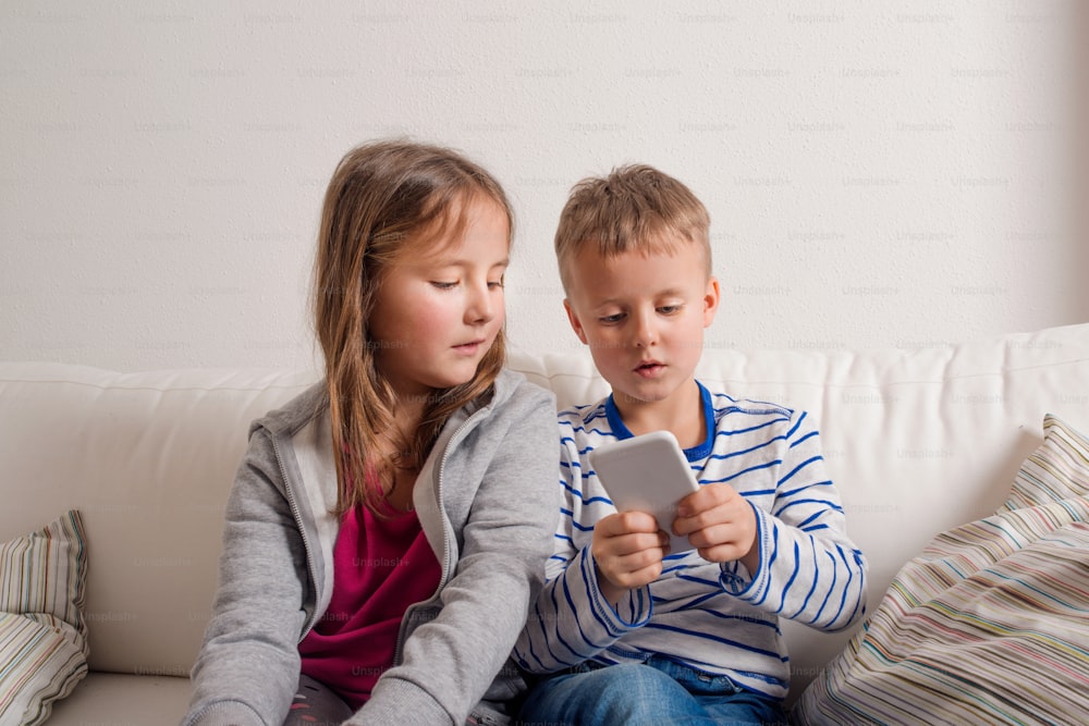 Little girl and boy sitting on sofa with a smart phone at home. Happy children playing indoors
