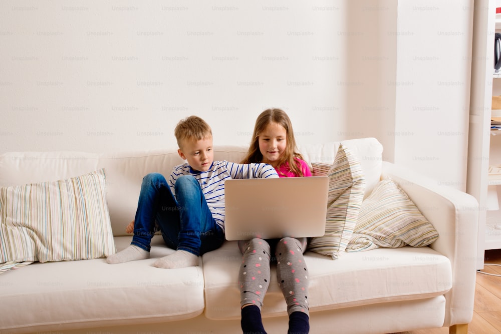 Niña y niño sentados en el sofá con una computadora portátil en casa. Niños felices jugando en el interior usando la PC.