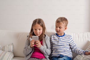 Little girl and boy sitting on sofa with a smart phone at home. Happy children playing indoors, copy space