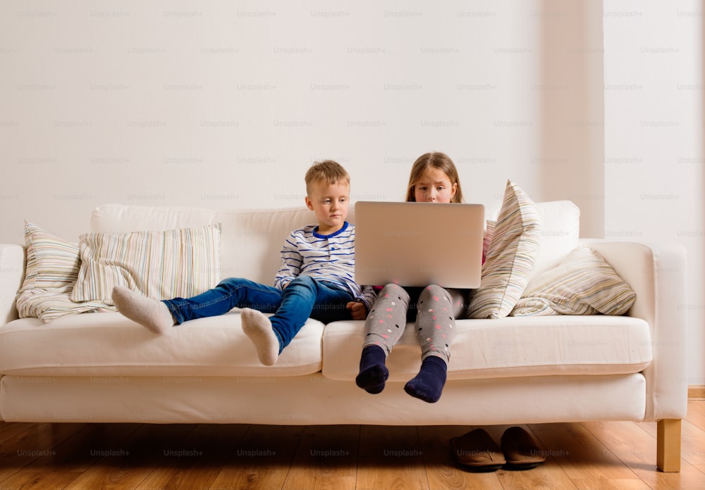 Petite fille et garçon assis sur un canapé avec un ordinateur portable à la maison. Des enfants heureux jouant à l’intérieur à l’aide d’un PC.