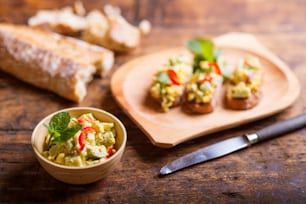 Spread made of avocado and other ingredients in a bowl and on slices of bruschetta. Studio shot on old wooden table background