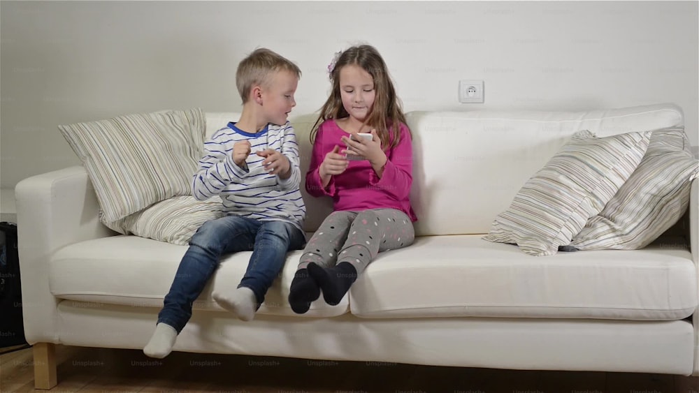 Little girl and boy sitting on sofa with a smart phone at home. Happy children playing indoors.