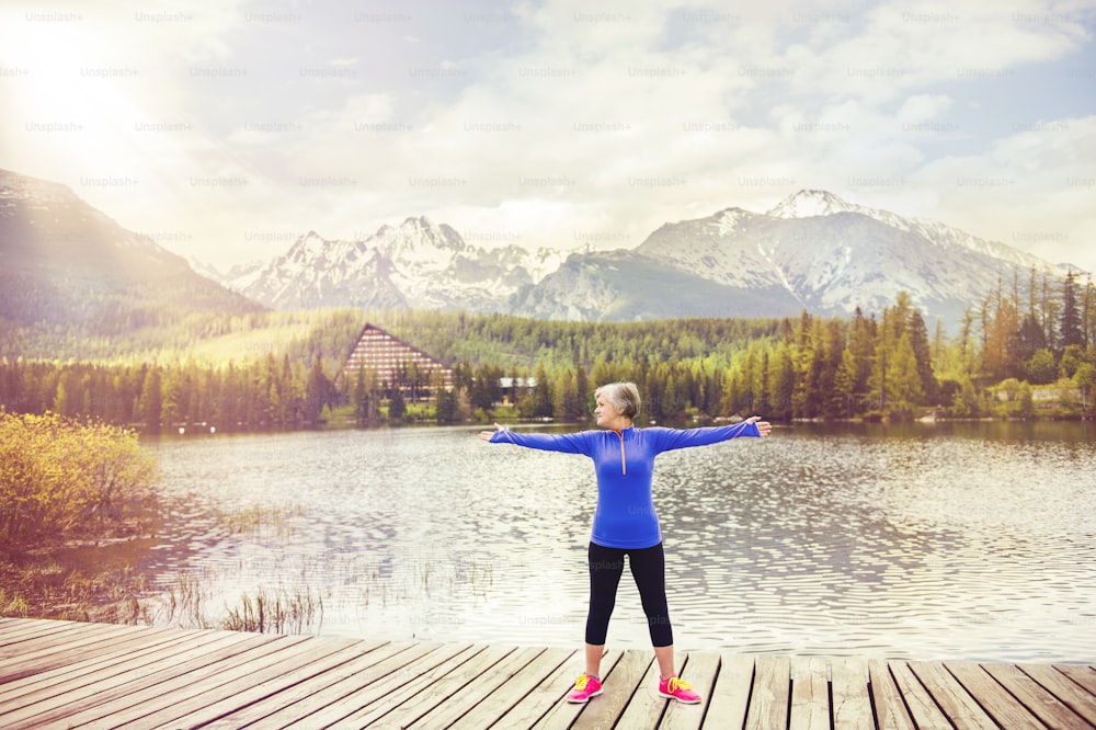 Senior woman is exercising on the pier by the tarn in beautiful mountains, hills and hotel in background