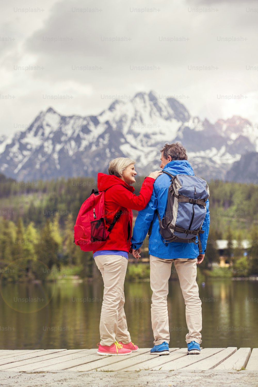 Coppia di escursionisti anziani durante la passeggiata intorno al laghetto in bellissime montagne, colline e hotel sullo sfondo