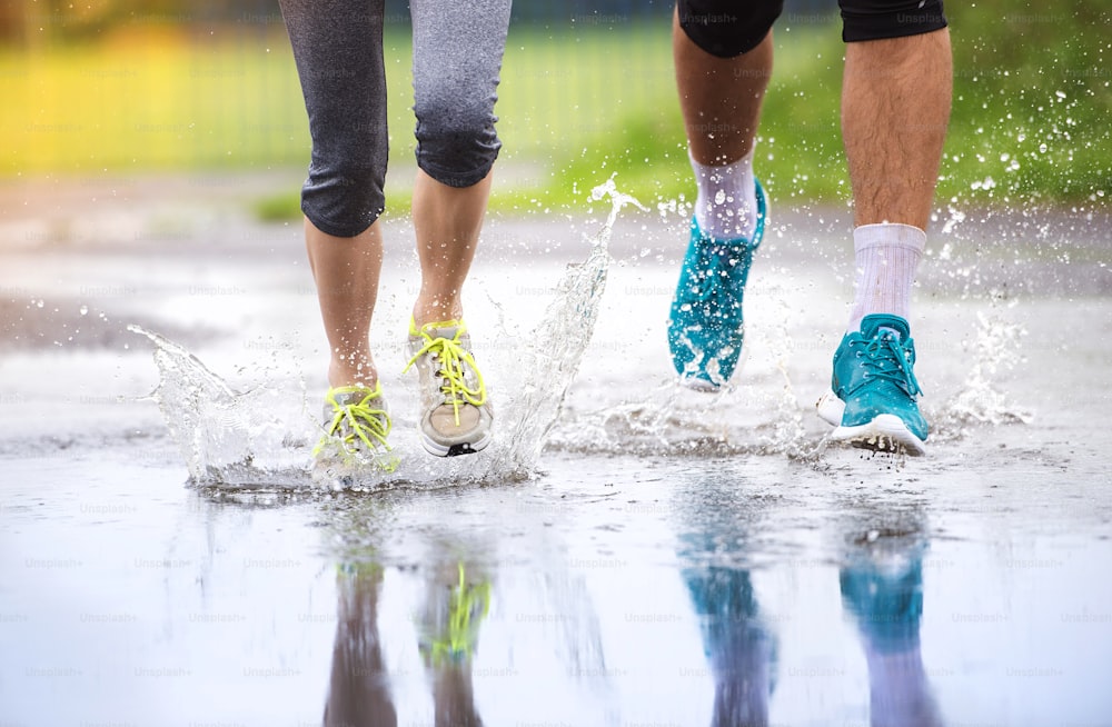 Young couple running on asphalt sports field in rainy weather. Details of legs and sports shoes splashing in puddles.