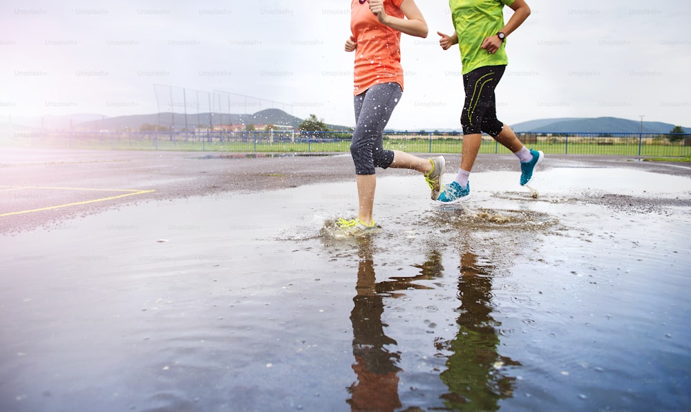 Giovane coppia che corre sul campo sportivo asfaltato in tempo piovoso. Dettagli di gambe e scarpe sportive che schizzano nelle pozzanghere.