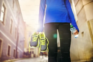 Unrecognizable female runner carrying her running shoes and bottle of water in old city center