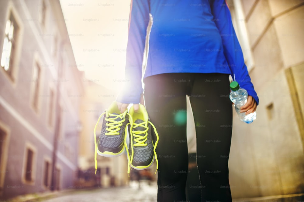 Unrecognizable female runner carrying her running shoes and bottle of water in old city center