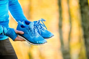 Unrecognizable runner in blue sweatshirt holding pair of sports shoes outside in colorful sunny autumn nature.