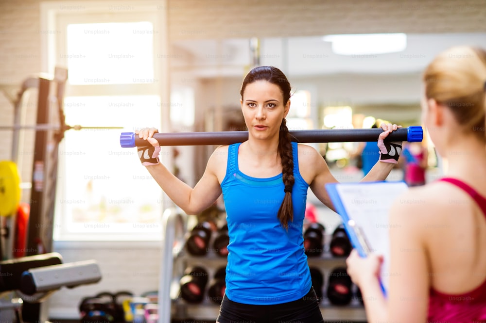 Mujer en camiseta azul haciendo ejercicio con barra en un gimnasio con su entrenador personal escribiendo en el plan de ejercicios en el portapapeles