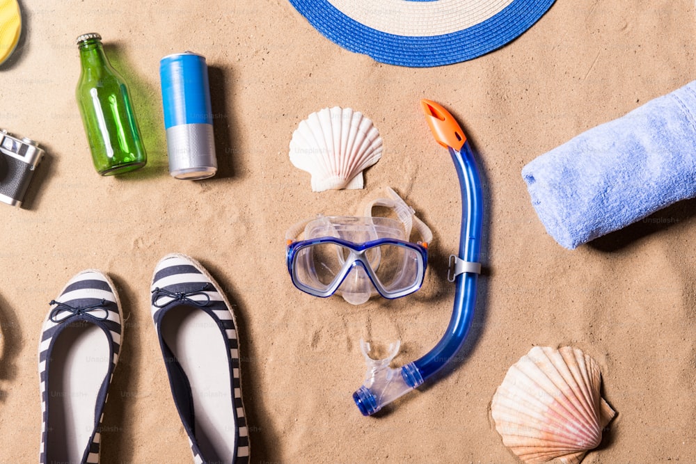 Composizione per le vacanze estive con occhiali, scarpe, conchiglia, asciugamano, bottiglia, lattina e altre cose su una spiaggia. Sfondo di sabbia, scatto in studio, flat lay.