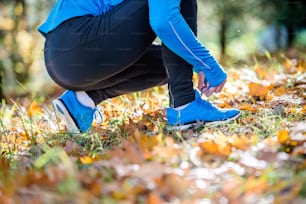 Unrecognizable runner in blue sweatshirt outside in colorful sunny autumn nature sitting on the ground, tying shoelaces