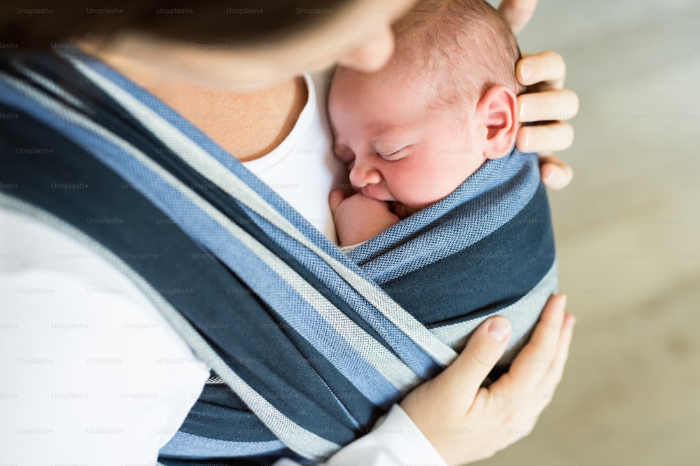 Primer plano de una madre joven irreconocible con su hijo recién nacido en cabestrillo en casa