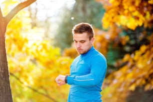 Joven y guapo corredor hipster en sudadera azul midiendo el tiempo con su reloj afuera en la colorida naturaleza soleada del otoño