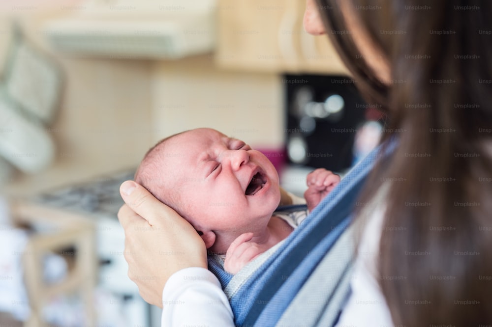 Primer plano de una madre joven irreconocible con su hijo recién nacido llorando en cabestrillo en casa