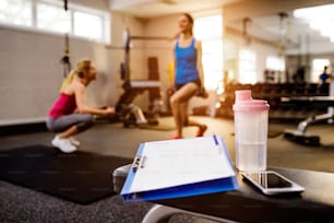 Gym close up, woman with personal trainer exercising squats, clipboard with work out plan, water bottle, smart phone
