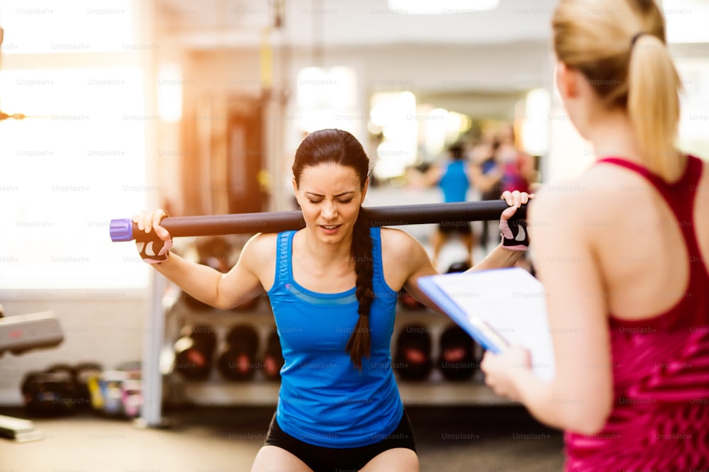 Primer plano de una atractiva mujer en forma en camiseta azul haciendo ejercicio en un gimnasio con su entrenador personal escribiendo en el plan de ejercicios en el portapapeles