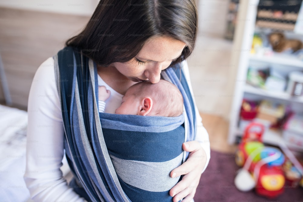 Primer plano de una hermosa madre joven con su hijo recién nacido en cabestrillo en casa, besándolo
