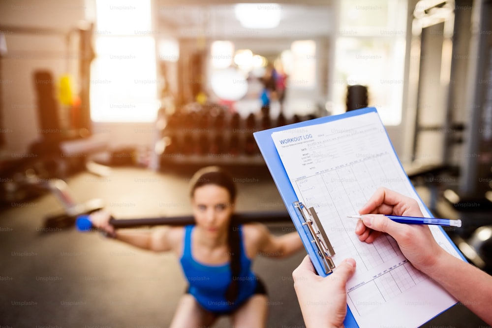 Close up of an attractive fit woman in blue singlet exercising in a gym with her personal trainer writing into exercise plan on clipboard